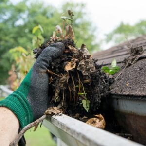 how to prepare your house for winter - person with glove cleaning debris out of clogged rain gutter